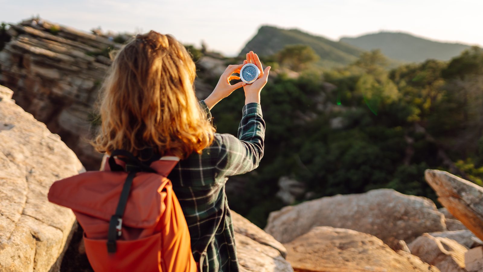 A walker with a red rucksack holding a compass up against a hazy, hilly view