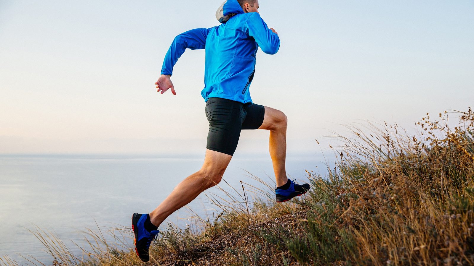 A runner in a blue jacket climbing a steep grassy hillside above the sea